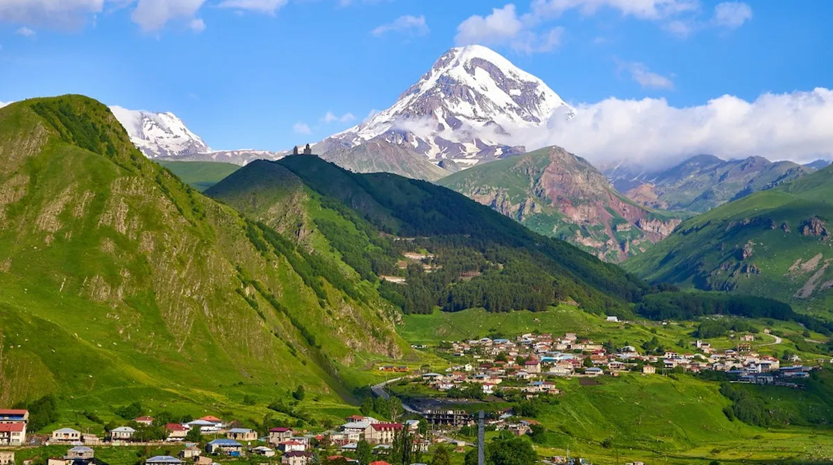 Main photo for Kazbegi private tour, showcasing the village of Stepantsminda in the lush green valley with the towering, snow-capped Mount Kazbek and the iconic Gergeti Trinity Church perched on the ridge under a blue sky